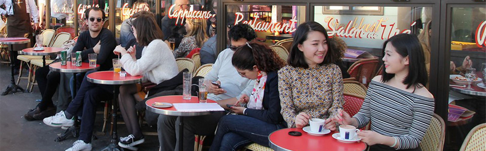 People are sitting at small tables outside of a cafe. They are looking at each other and chatting.